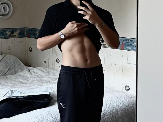 CharliePassion, a lean, muscular guy, lifts his black shirt to reveal a defined six-pack, wearing black pants, holds a phone, watch on wrist, standing in a bedroom with a messy bed.