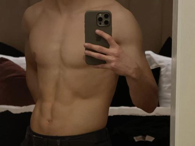 Photograph of a muscular, shirtless CharliePassion taking a mirror selfie with a smartphone, showcasing defined abs and chest, wearing dark pants, slight shadowing, and a bedroom setting.
