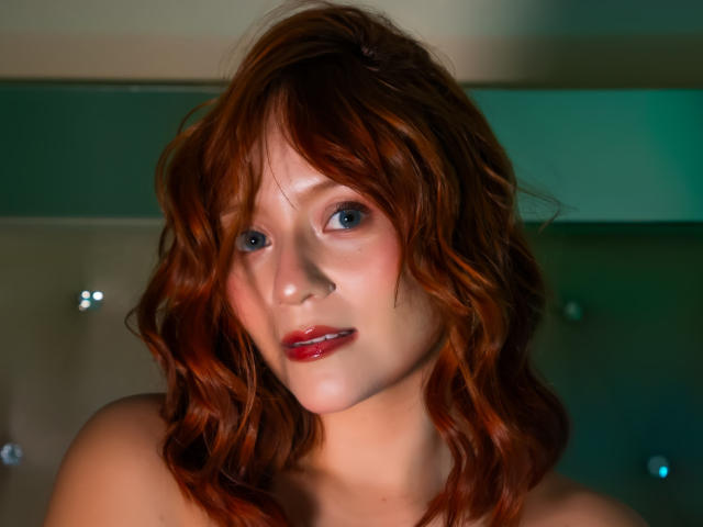Close-up photograph of EmilyKlein with wavy auburn hair, blue eyes, and red lipstick. Her expression is slightly seductive, with soft shadows highlighting her facial features. Lighting accentuates her skin's smooth texture.