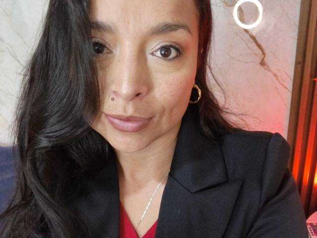 Close-up of StangelarRivera, a Latina woman with long black hair, wearing a black blazer over a red shirt, hoop earrings, and a nose piercing. Red light in background. Focused gaze.