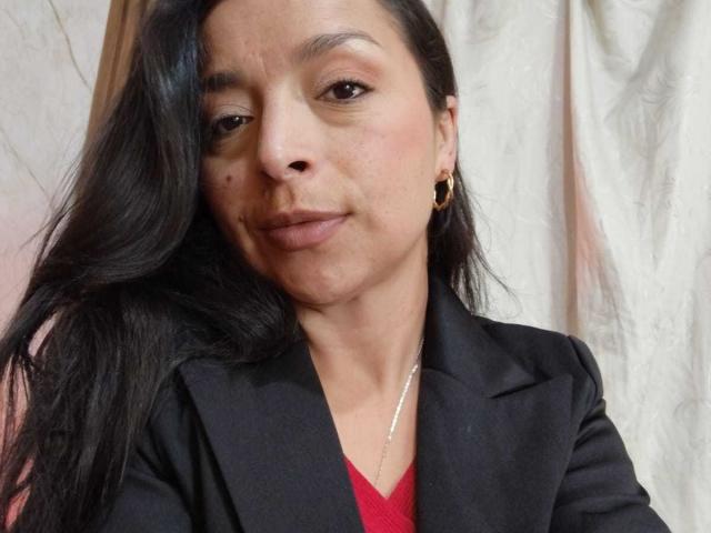 Close-up of StangelarRivera, a woman with long black hair, wearing a black blazer over a red top, gold hoop earrings, and a thin necklace. She has a neutral expression, some acne.