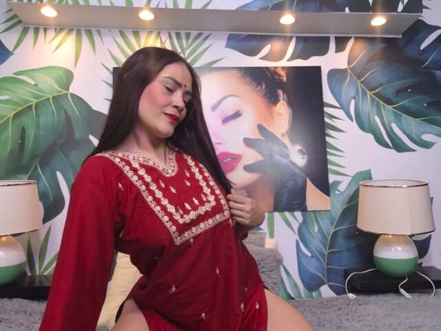 ShantalaKrisha, con cabello largo y oscuro, vestida con un vestido rojo con bordados dorados, sentada en el suelo, con una mano en el pecho, atractiva. Fondo de mural tropical, dos lámparas, iluminación suave, expresión sensual..