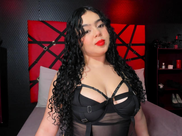 AprielKeith, curvy Latina with long black curly hair, wearing a sheer black bra with straps and buckles, red lips, centered in a red and black geometric headboard bedroom. Fucking sexy.