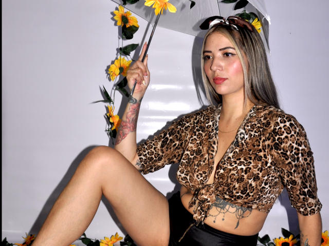 CoralRosse zit met gespreide benen, een witte paraplu met gele bloemen vasthoudend. Een topje met luipaardprint, zwarte korte broek. Getatoeëerde arm, donker steil haar, intense blik. Heel aantrekkelijk..
