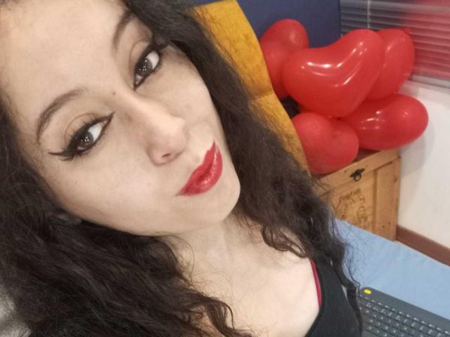 Close-up of KendraSimons with pale skin, black curly hair, and red lipstick, gazing seductively at the camera. Red inflatable dildos in background, sexual tension, fucking sexy look.