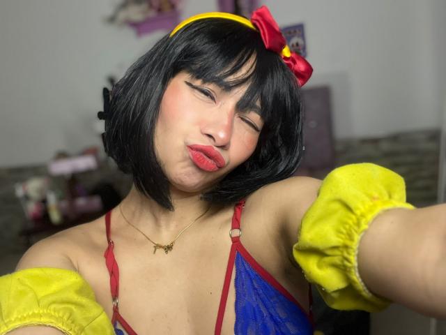 MelannyTaylorr, cosplaying as Snow White, with black bob haircut, red bow, yellow headband, red and blue top, yellow puffed sleeves. Pouting lips, red lipstick, slight smile, sexy pose, blurred background.
