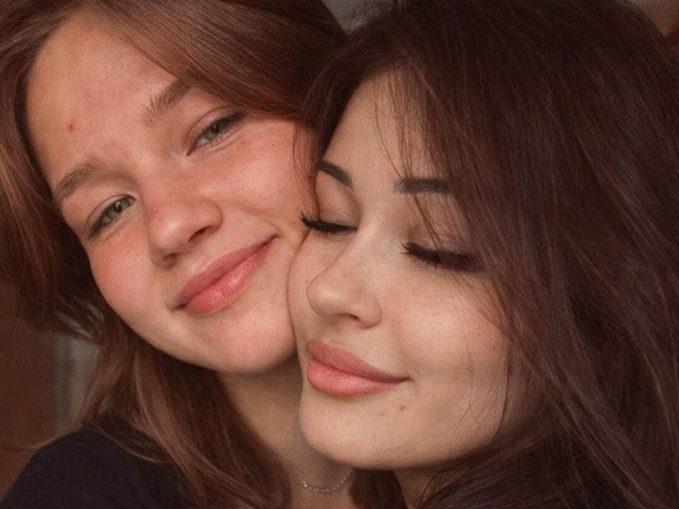 Gros plan de deux femmes à la peau claire et aux cheveux bruns, HelliForJeren à gauche, souriante, son visage pressé contre visage les yeux fermés de HelliForJeren sur la droite, tous deux avec des lèvres roses..