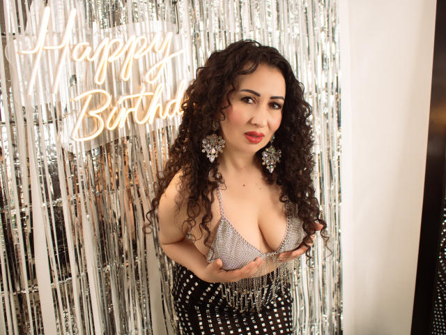 SamanthaAntolin stands against a silver curtain, wearing a sparkly, low-cut bikini top that reveals large breasts. She has long, curly black hair and red lipstick. Neon text "Happy Birthday" glows behind her.