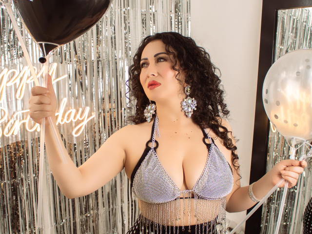 SamanthaAntolin, with curly black hair, wearing a sparkly white bikini top, holding a balloon and a black curtain. Silver tinsel background. Bright lights, sexy, confident pose, large breasts.