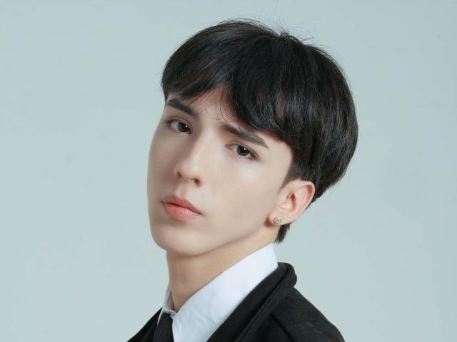 Close-up photo of AerumChaee with short black hair, pale skin, and a serious expression. Wearing a white shirt and black jacket. Background is plain white. Sexual tension from direct gaze.
