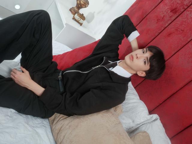 AerumChaee lag auf einem Bett, die Beine gespreizt, und trug einen schwarzen Anzug mit weißem Hemd, wobei Kopfhörer herunterhingen. Roter Kopfteil, weiße Kissen, beiges Kissen. Entspannte, sexy Pose, leichtes Lächeln. Lässige, selbstbewusste Ausstrahlung.