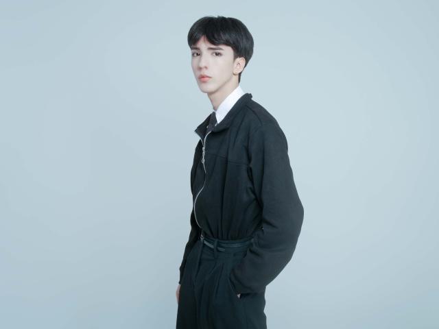 Foto van AerumChaee met kort zwart haar, gekleed in een zwarte jas en een wit shirt, staand voor een helder witte achtergrond. Achtergrond. Handen in de zakken, serieuze uitdrukking, subtiele seksuele spanning.