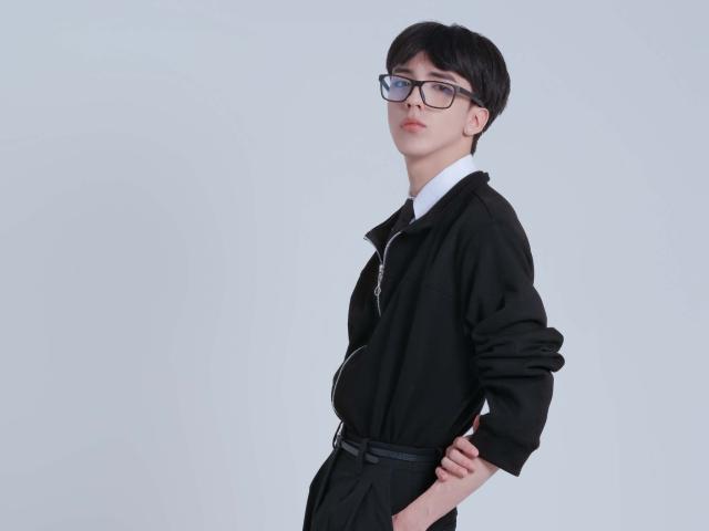 Fotografía de AerumChaee con cabello corto y negro, usando gafas negras y una camisa negra con cuello blanco, de pie. contra un fondo blanco. Manos en las caderas, un aire sexy, pero también un poco intelectual..