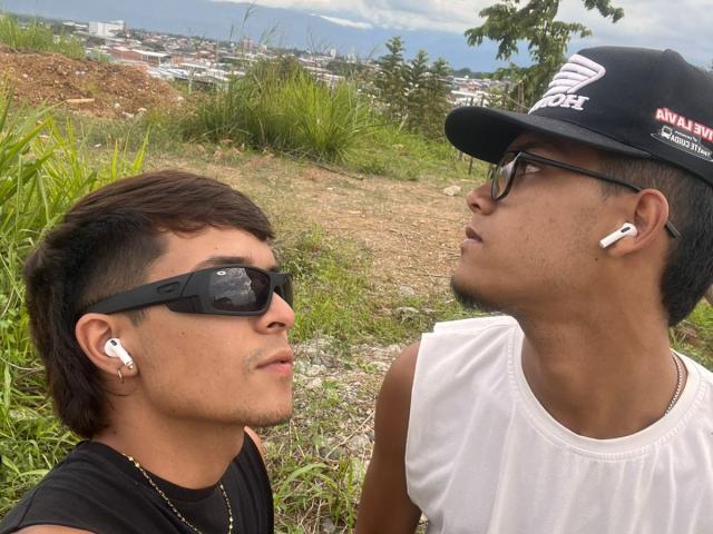 Two men with medium brown skin, DanAndKamadeva on the left in black t-shirt and sunglasses, DanAndKamadeva on the right in white tank top and black cap, grassy hillside, blue ocean. Earbuds, intense gaze.