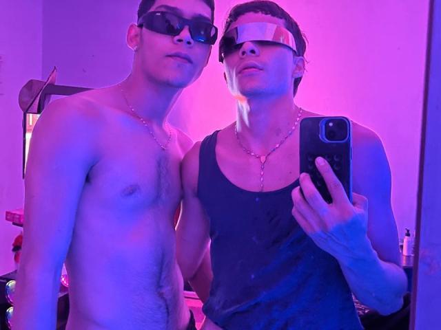 Dos hombres de pie, muy cerca uno del otro, ambos con gafas de sol y cadenas de oro. Uno sin camisa, el otro con una camiseta negra. Fondo de luz neón morada y rosa. Una persona tomando una selfie con el teléfono. Increíblemente atractiva..