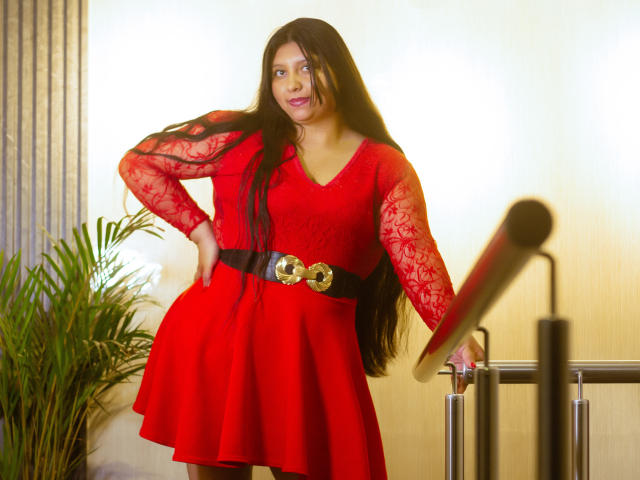 SophieGalli è in piedi, sicura di sé, con un abito rosso vivace in pizzo, una cintura nera e una fibbia dorata. Ha lunghi capelli neri, un sorriso seducente e appoggia una mano su una ringhiera..