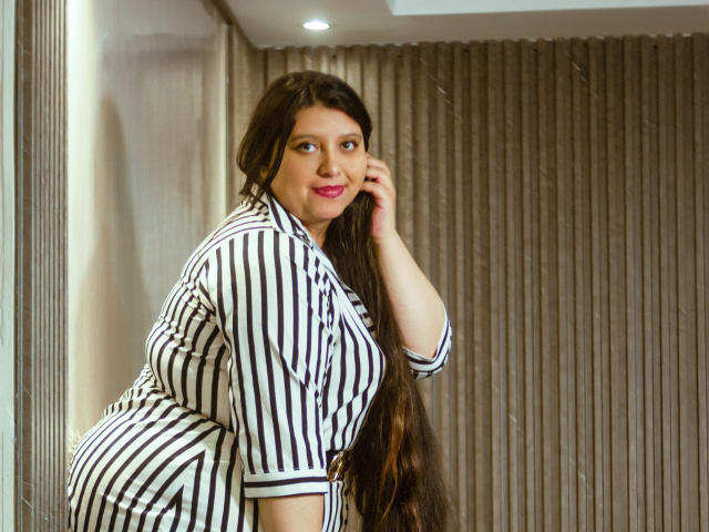 SophieGalli, volslank met grote borsten, draagt een zwart-wit gestreepte blouse en leunt tegen een muur, met haar hand in haar haar, lang donker haar, rode lippenstift, verleidelijke glimlach, grijze gordijnen op de achtergrond.