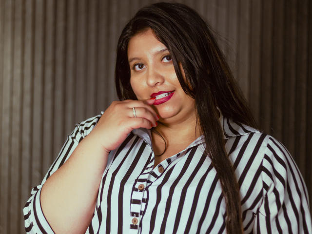 Fotografie von SophieGalli mit langen, schwarzen Haaren, die ein schwarz-weiß gestreiftes Hemd trägt und ihre rot geschminkten Lippen beißt. Nagel. Sie hat ein rundes Gesicht, braune Augen und einen verführerischen Blick..