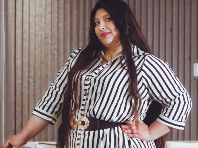 Fotografía de SophieGalli, una mujer de curvas con cabello largo y negro, que lleva una camisa a rayas blancas y negras y una falda negra ancha. cinturón con una hebilla decorativa grande. Ella está de pie con confianza, con una mano en la cadera, mirando directamente a la cámara. Sus labios están pintados de un rojo brillante.