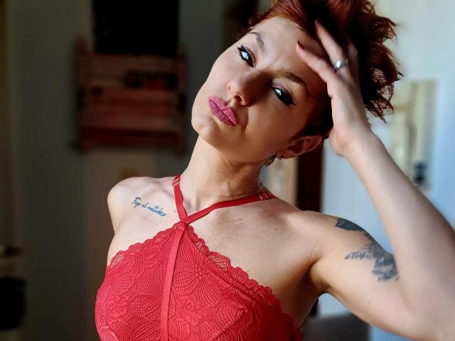 DreamityCamy, kurze rote Haare, Hand im Haar, rotes Spitzenoberteil, Tattoos an der Schulter, helle Haut, verführerischer Blick, markante Kieferpartie. leicht geöffneter Mund, weiches Licht, intensiver Ausdruck, sexuelle Spannung.