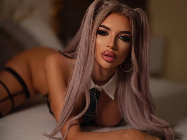 JuicyJessye, lying on a bed, face close to the camera, long platinum blonde hair, dark eye makeup, full pink lips, black strappy lingerie, seductive pose, soft lighting, sexy tension.