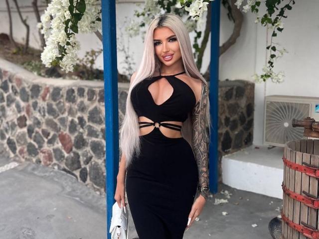 JuicyJessye, con lunghi capelli biondo platino, indossa un abito nero aderente con aperture, ed è in piedi tra pilastri blu. Fiori bianchi sullo sfondo, un muro di pietra e una botte a destra. Molto attraente..