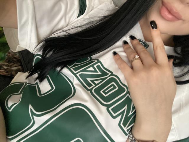 Gros plan d'Olieri portant un maillot de sport blanc et vert, avec des ongles noirs, touchant ses lèvres avec un doigt, et portant une bague noire. présente, cheveux longs et noirs, tension sexuelle, pose suggestive.