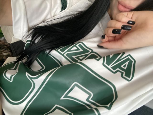 Close-up van Olieri met lang zwart haar, die een witte sporttrui met groene letters draagt en de stof vasthoudt. Zwart geverfde nagels, penis zichtbaar onder het shirt, sexy spanning..