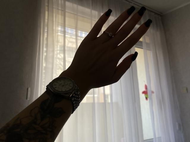 Silhouette of Olieri's hand with black nail polish, wearing a silver watch and ring, raised palm facing forward. Soft light from window in background. Fingers spread, tense, slightly provocative.