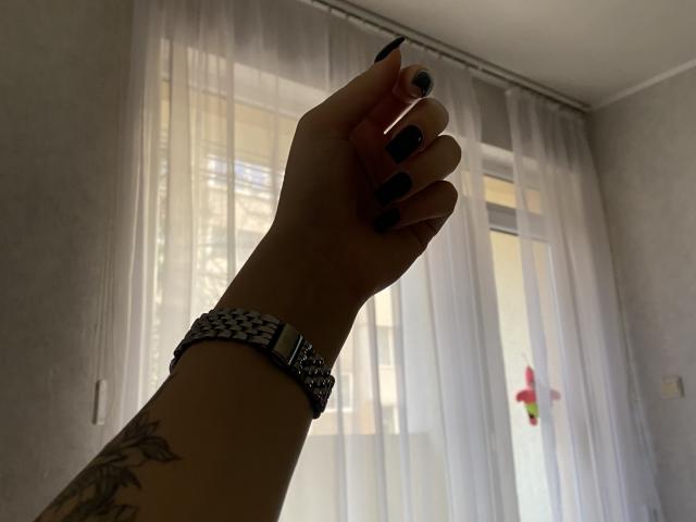 Silhouette de la main d'Olieri, avec du vernis à ongles noir, portant une montre en argent, les doigts levés vers des rideaux transparents. Tatouage visible sur l'avant-bras. La lumière du soleil filtre à travers les fenêtres. Tension sexuelle due à une pose suggestive..