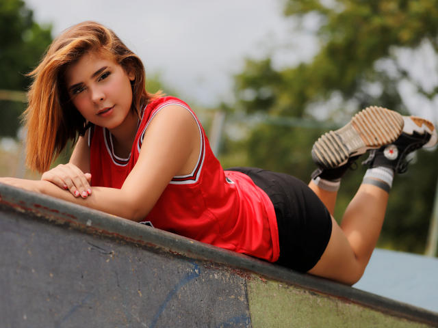 Zara Vidal liegt auf einem Betonvorsprung, die Beine angewinkelt, und trägt ein rotes Top ohne Ärmel, schwarze Shorts und schwarz sneakers. Sie hat braunes Haar, einen verführerischen Look und ein betörendes Lächeln..