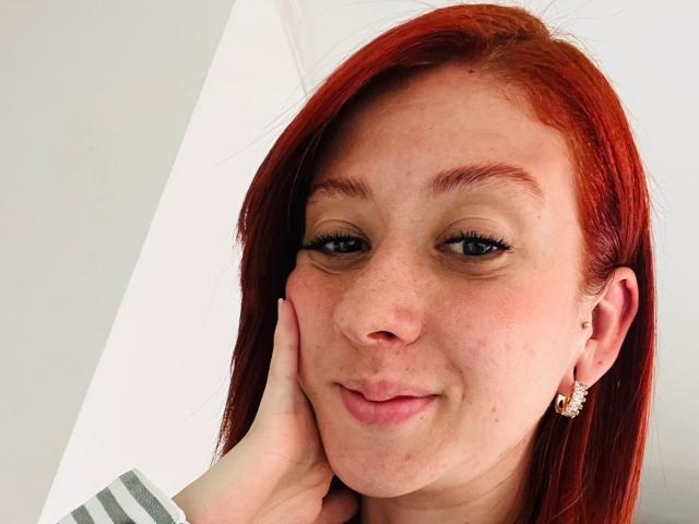 Foto ravvicinata di Valeriaaf, una donna dai capelli rossi con lentiggini, che tocca leggermente la guancia con la mano e sorride. leggermente, con piccoli orecchini a forma di stella, sfondo bianco brillante. Che carino!.