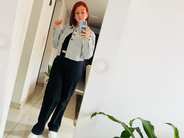 Valeriaaf stands in a brightly lit room, taking a mirror selfie. She's wearing a striped shirt, black pants, and a black tie. Her red hair contrasts with the white background. She's holding a phone in her right hand.