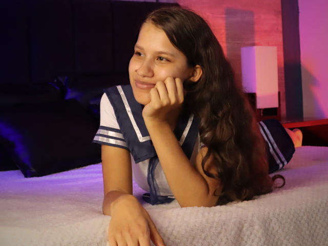 Emma, una joven con cabello largo y oscuro, está acostada en una cama, vestida con un uniforme de marinera azul marino y blanco, y sonríe. de manera seductora, su mano tocando su mejilla, iluminación rosa y azul neón..