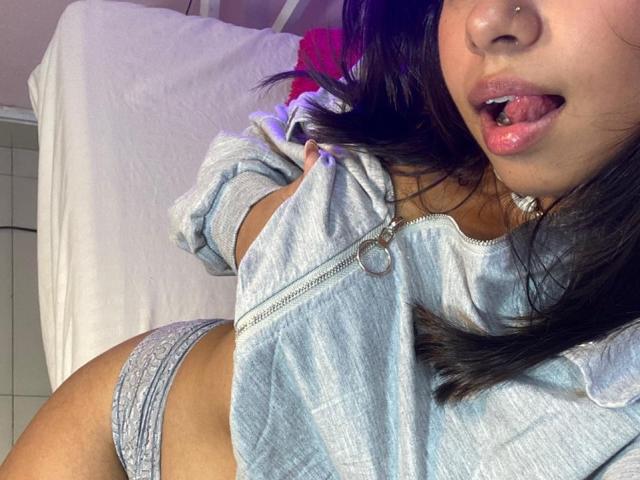 Close-up of CelesteMorel with her tongue out, wearing a silver zip-up top and gray lace panties. Her black hair is slightly messy, and there's a pink flower in the background. Fucking sexy.
