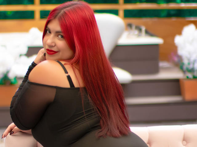 AmberCutel mit leuchtend rotem Haar, die verschmitzt lächelt, lehnt sich vor. Sie trägt ein schwarzes Top mit einem Schulterteil, hat große Brüste, dicke Oberschenkel und sieht sexy aus. Pose, unscharfer Hintergrund mit leuchtenden Lichtern, verführerisch, lebendige Farben, selbstbewusster Ausdruck..