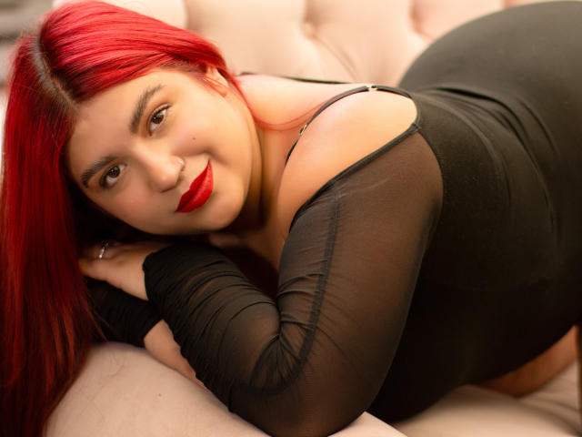 AmberCutel, con i suoi capelli rosso acceso, è sdraiata su un letto, con la testa rivolta verso la telecamera, indossando una maglia nera trasparente che lascia intravedere... spalla e lato del seno, rossetto rosso, espressione seducente, tessuto nero.