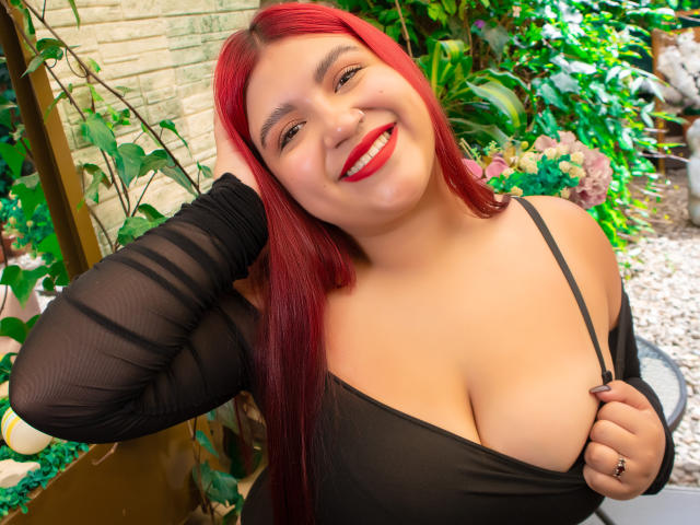 AmberCutel, with long red hair, smiling widely, pulls down black top to reveal large breasts. Wearing black sleeves, red lipstick, bright red nails, leaning against wooden structure, green plants in background.