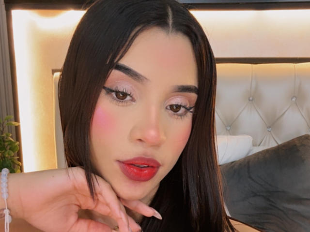 Close-up photo of LunaVali, a sexy woman with long black hair, red lipstick, and pink blush. She gazes seductively at the camera with her hand on her chin. Soft lighting.