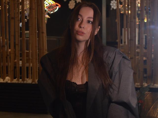 Murris, avec ses longs cheveux bruns, porte une veste grise par-dessus un haut noir en dentelle, et est assise dans une pièce faiblement éclairée avec des panneaux en bambou. L'éclairage doux met en valeur ses traits, créant une ambiance sensuelle..