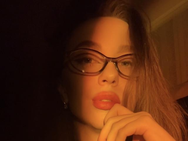 Close-up of Murris with sultry expression, biting finger, large red lips, dark brown hair, and black-rimmed glasses. Warm yellow-orange lighting creates a sexy, intimate atmosphere.
