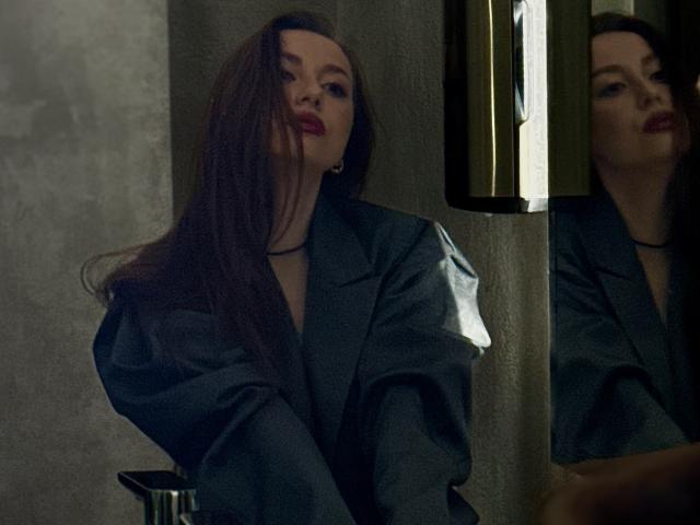 Photographie sombre de Murris, avec de longs cheveux bruns, portant une veste grise, assise dans une pièce sombre, avec un reflet dans un miroir. regard droit, regard intense, lèvres rouges, éclairage sombre, expression sérieuse.