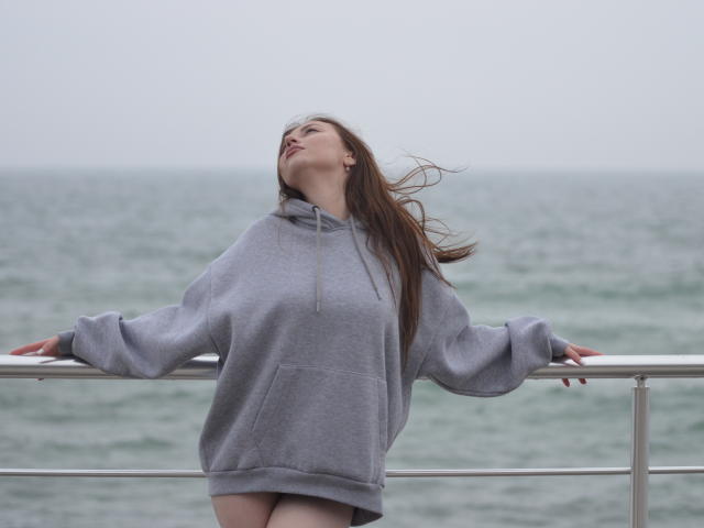 Murris se tient sur une rambarde en métal près de l'océan, la tête penchée en arrière, ses longs cheveux bruns flottant au vent. Portant un sweat à capuche gris ample, les mains agrippant la rambarde. La mer et le ciel se fondent en arrière-plan..