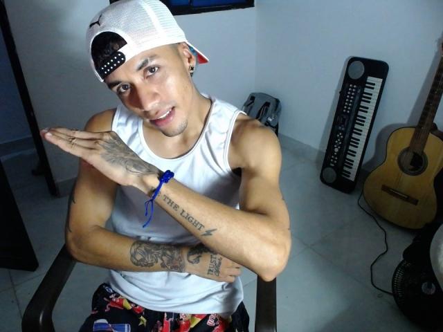 JersiSexlov, sin camisa, gorra blanca puesta al revés, camiseta blanca sin mangas, tatuajes en los brazos, brazos cruzados, sonriendo, pantalones de colores. foto digital, teclado, guitarra, iluminación brillante, pose informal, tatuajes visibles, ambiente relajado..