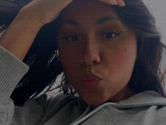 Close-up of AnaRome, dark-haired woman with a nose ring, pouting lips, and hand in hair. Wearing a gray hoodie. Shadowy lighting, intense gaze, slightly blurred texture. Fucking sexy.