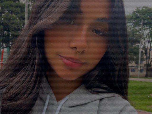 Close-up of AnaRome, a young woman with long, dark brown hair, brown skin, and a gold nose ring. She's wearing a gray hoodie, slightly smiling, with visible outdoor park background.