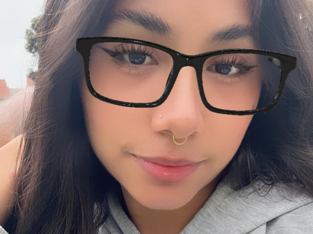 Primer plano de AnaRome, una joven con cabello largo y oscuro, gafas de montura negra y un piercing dorado en la nariz, mirando directamente a la cámara, con una ligera sonrisa. Ligera tensión sexual.