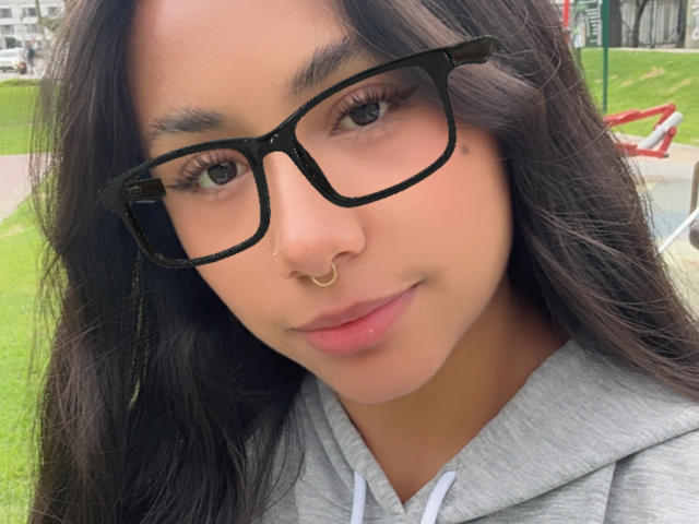 Close-up de AnaRome, uma jovem com cabelos longos e pretos, óculos de aro preto e um piercing de ouro no nariz. Ela está vestindo um moletom cinza, olhando diretamente para a câmera com um leve sorriso. Fundo de parque ao ar livre..