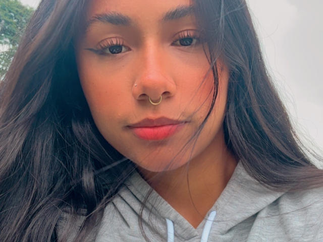 Close-up foto van AnaRome met lang zwart haar, die een grijze hoodie draagt, een neusring heeft en rode lipstick, en recht in de camera kijkt. De camera, met de lippen lichtjes open. Zacht licht, lichte bries in het haar..