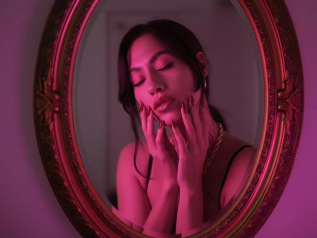 Photographie de MelisaDaltonn reflétée dans un miroir ovale orné, baignée d'une lumière rose et violette. Elle a les yeux fermés, les mains posées sur ses joues, et elle porte un haut noir à bretelles. Tension sexuelle.